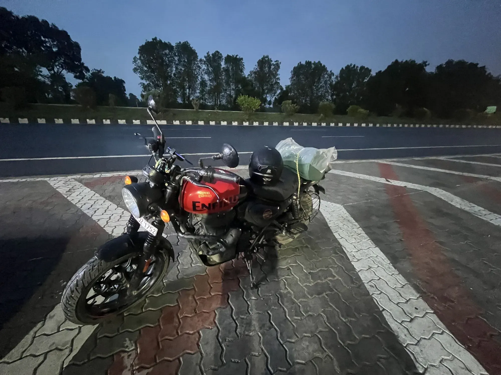 The red Royal Enfield Hunter 350 parked at a highway rest stop at dusk, a helmet and a luggage bag strapped to the seat, with the phone mount on the handlebar